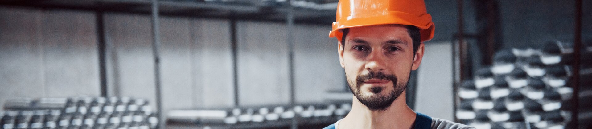 Industriearbeiter mit Schutzhelm und Handschuhen steht in Werkhalle mit Metallteilen