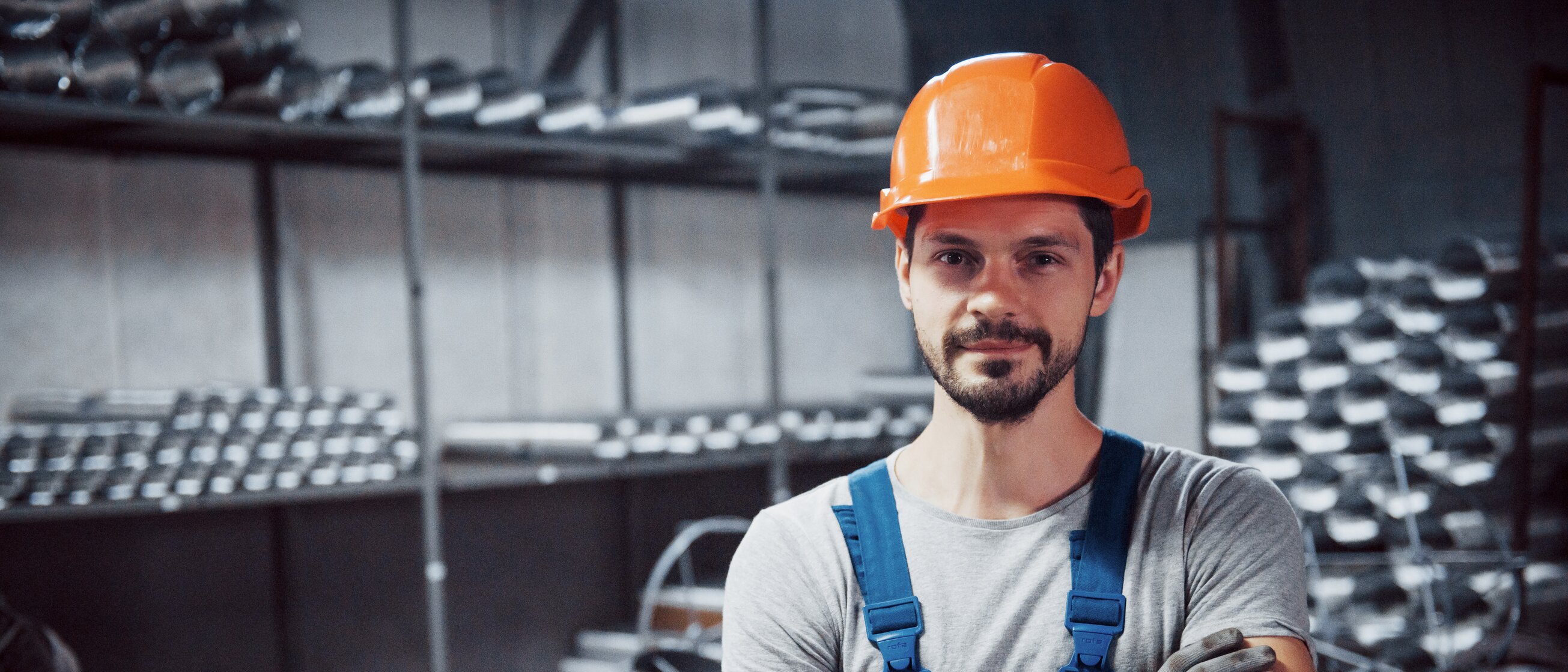 Industriearbeiter mit Schutzhelm und Handschuhen steht in Werkhalle mit Metallteilen