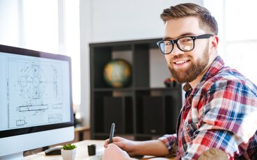 Lächelnder Mann mit Brille sitzt am Schreibtisch vor einem Computer mit technischer Zeichnung – Symbolbild für die Weiterbildung KI-Manager/-in IHK.