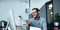 Mitarbeiter im Büro führt ein fröhliches Online-Meeting mit Headset am Computer durch.