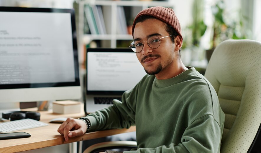 Junger Entwickler sitzt entspannt am Schreibtisch vor Monitoren mit Code und arbeitet an einem Softwareprojekt.