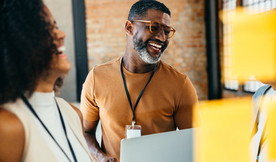 Teammeeting mit lächelndem Mitarbeiter, der einen Laptop hält und sich mit Kollegen austauscht.
