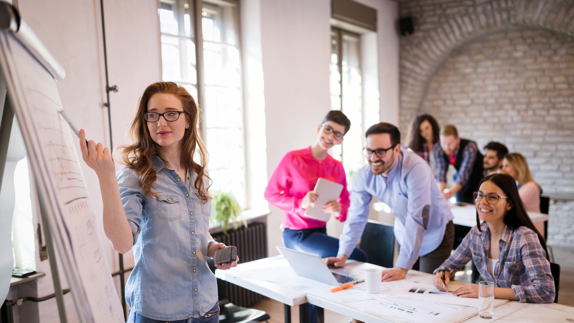 Junge Frau präsentiert Ergebnisse am Flipchart in einem modernen Seminarraum, während ein Team aufmerksam zuhört.