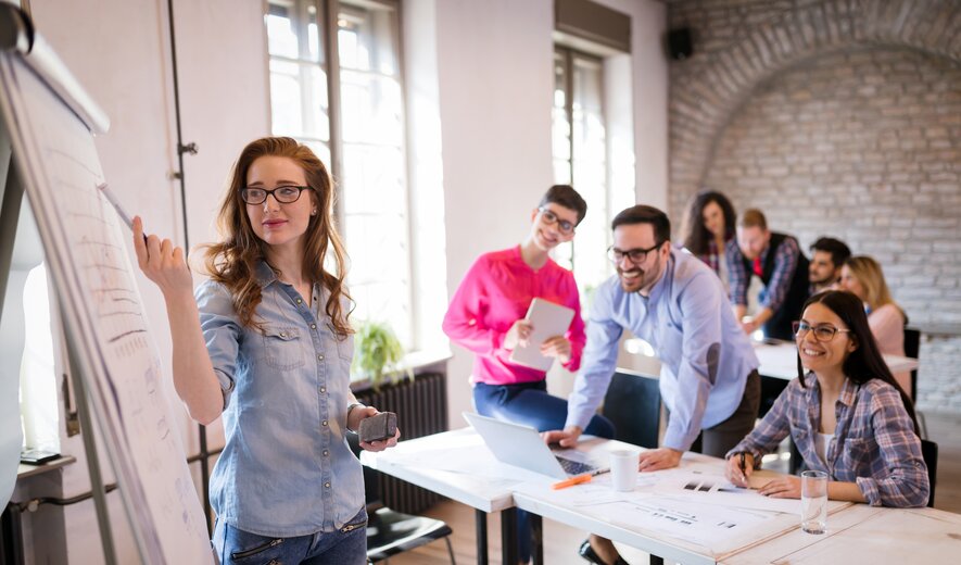 Junge Frau präsentiert Ergebnisse am Flipchart in einem modernen Seminarraum, während ein Team aufmerksam zuhört.