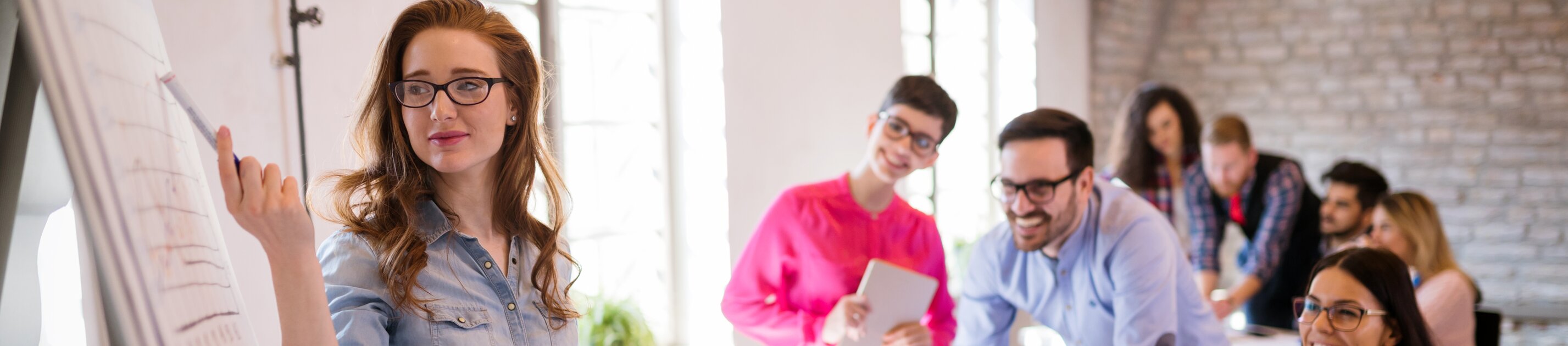 Junge Frau präsentiert Ergebnisse am Flipchart in einem modernen Seminarraum, während ein Team aufmerksam zuhört.