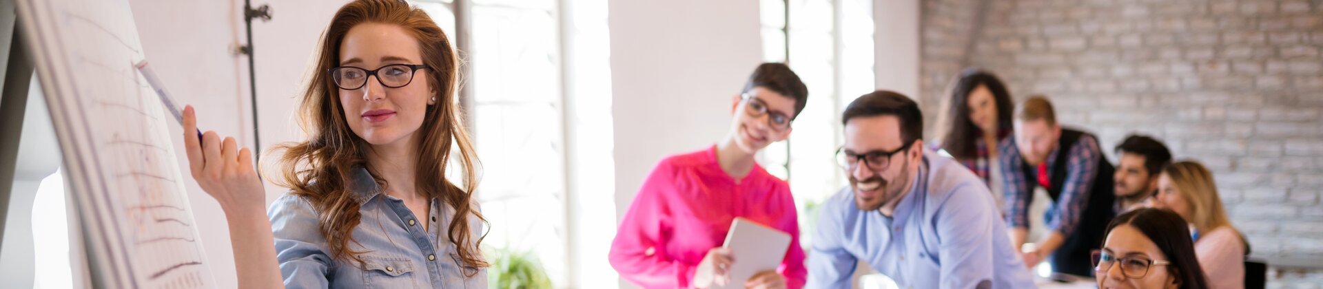 Junge Frau präsentiert Ergebnisse am Flipchart in einem modernen Seminarraum, während ein Team aufmerksam zuhört.