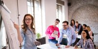 Junge Frau präsentiert Ergebnisse am Flipchart in einem modernen Seminarraum, während ein Team aufmerksam zuhört.