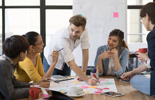 Weiterbildung im Projektmanagement: Eine Gruppe von Projektmitarbeitenden sitzt an einem Tisch und ein junger Mann schreibt etwas auf einen Post-It, der auf dem Tisch liegt. 