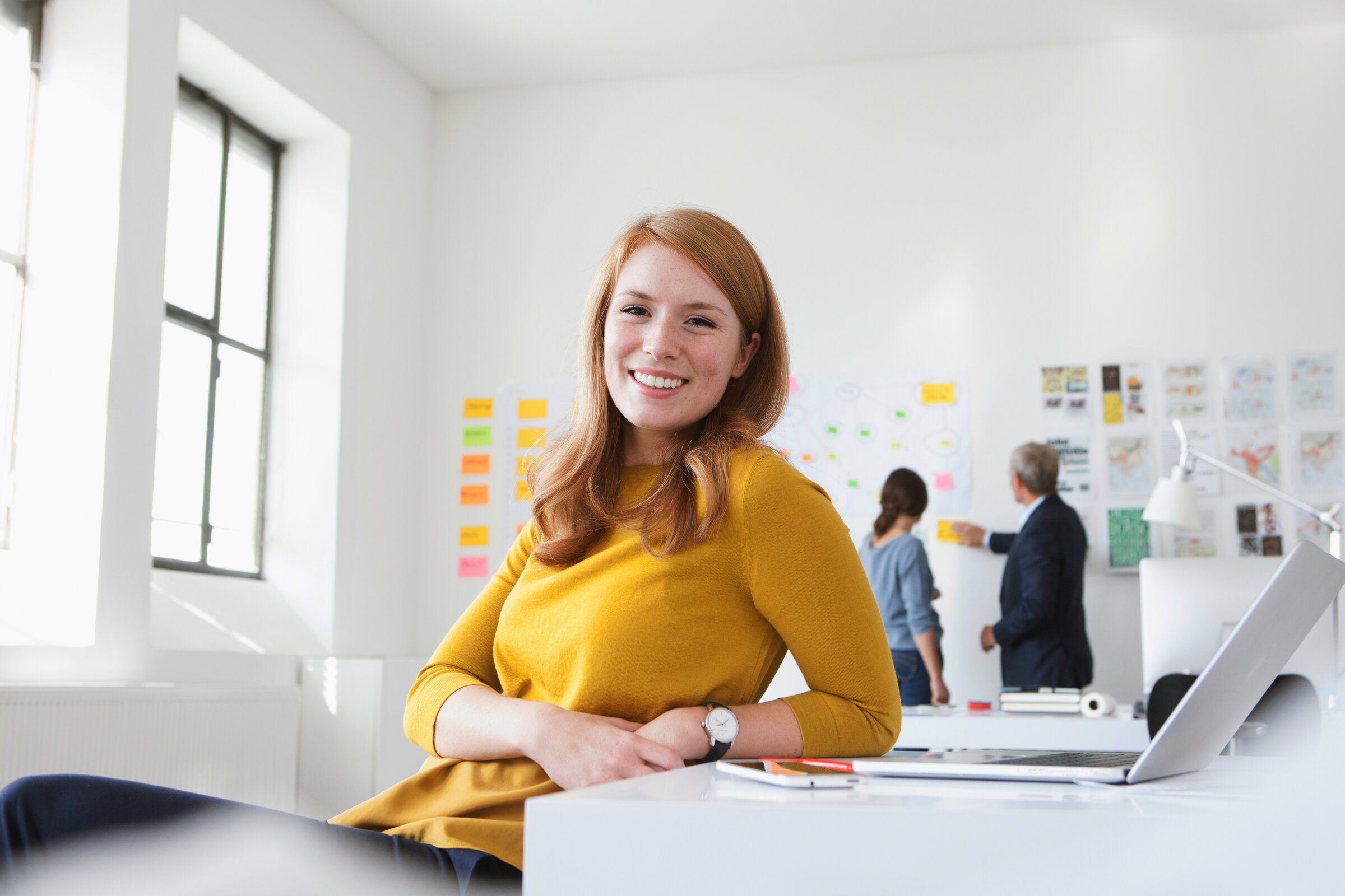 Weiterbildung im Projektmanagement: Junge Dame lehnt sich mit dem Ellbogen auf den Schreibtisch und schaut in die Kamera. 