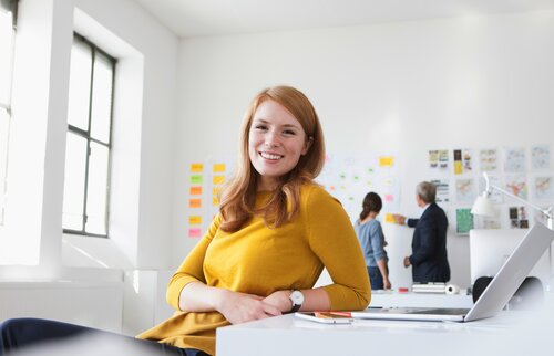 Weiterbildung als Projektmanager/-in IHK. Junge Dame sitzt vor ihrem Laptop in einem Meetingraum mit zwei weiteren Personen im Hintergrund. 