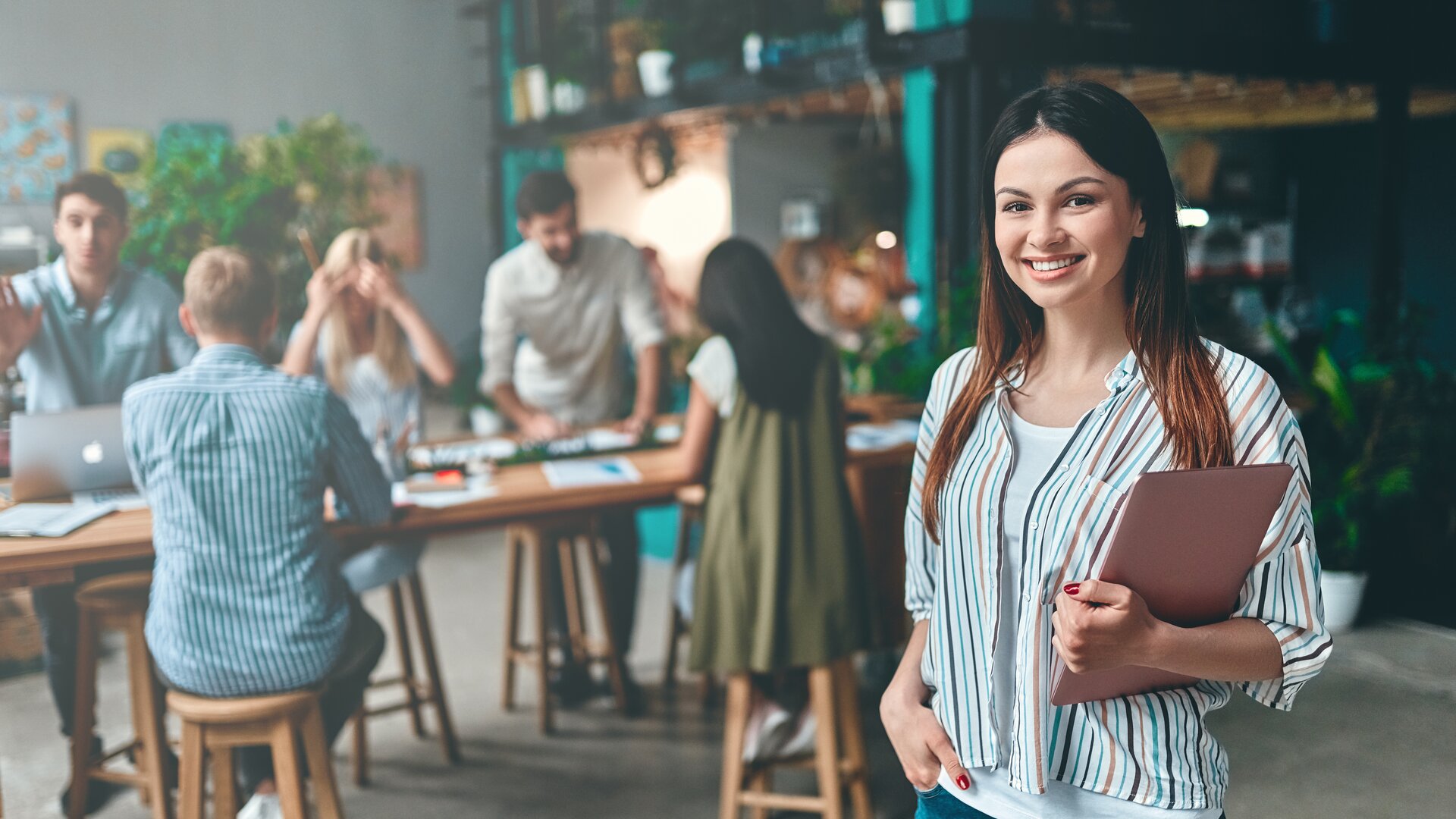Weiterbildung im Projektmanagement: Junge Dame steht im Vordergrund eines Raumes und schaut direkt in die Kamera. 