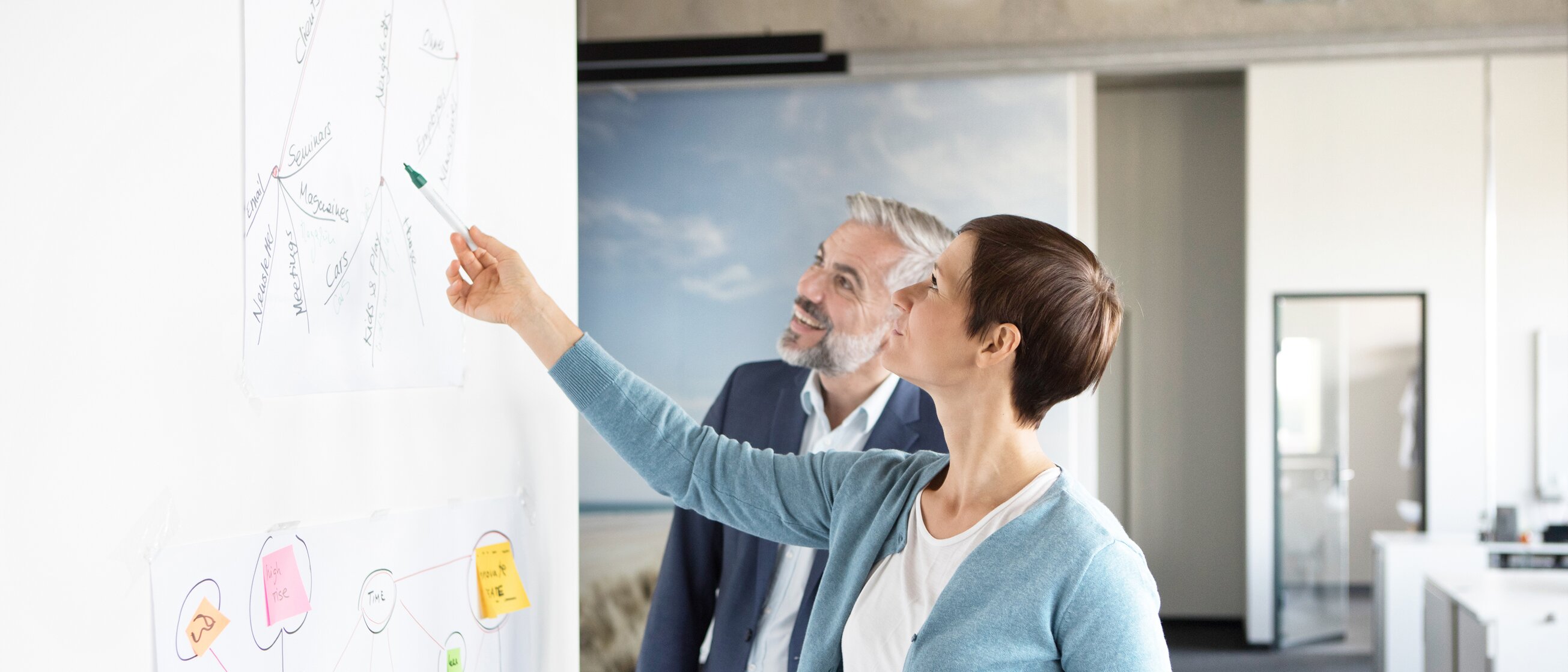 Weiterbildung im Projektmanagement: Eine Frau zeigt einem Mann etwas auf einer großen Projektmanagement-Chart.