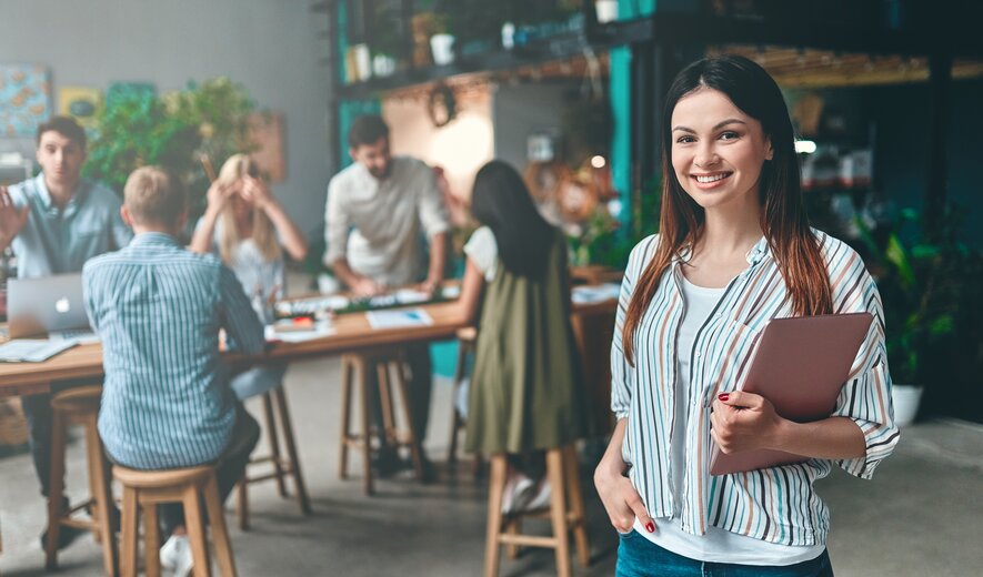 Weiterbildung im Projektmanagement: Junge Dame steht im Vordergrund eines Raumes und schaut direkt in die Kamera. 