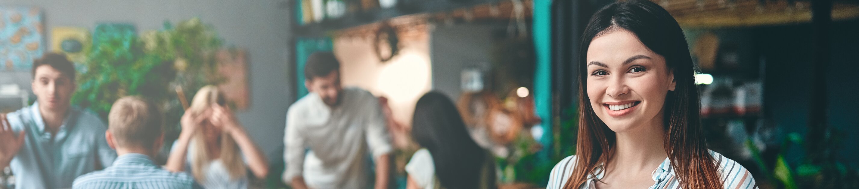 Weiterbildung im Projektmanagement: Junge Dame steht im Vordergrund eines Raumes und schaut direkt in die Kamera. 