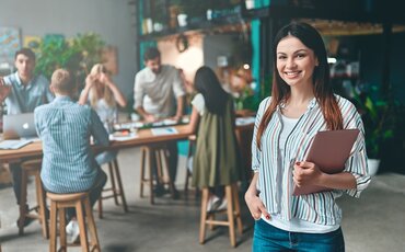 Junge Projektmanagerin präsentiert ein Konzept in einem modernen Büro, während ihr Team im Hintergrund an Projektstrategien arbeitet – Weiterbildung im Projektmanagement an der IHK Akademie.