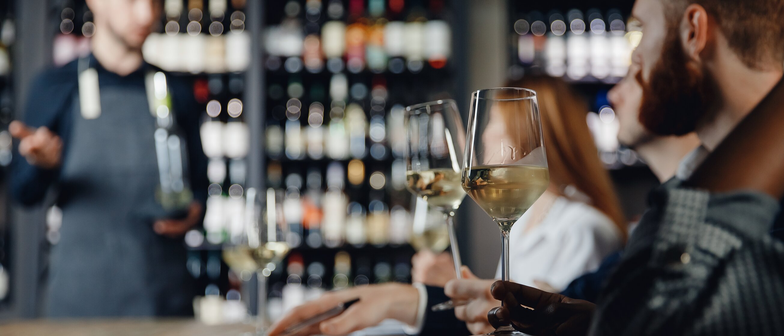 Weinverkostung in moderner Vinothek, Teilnehmende mit Weißweingläsern hören einem Sommelier aufmerksam zu