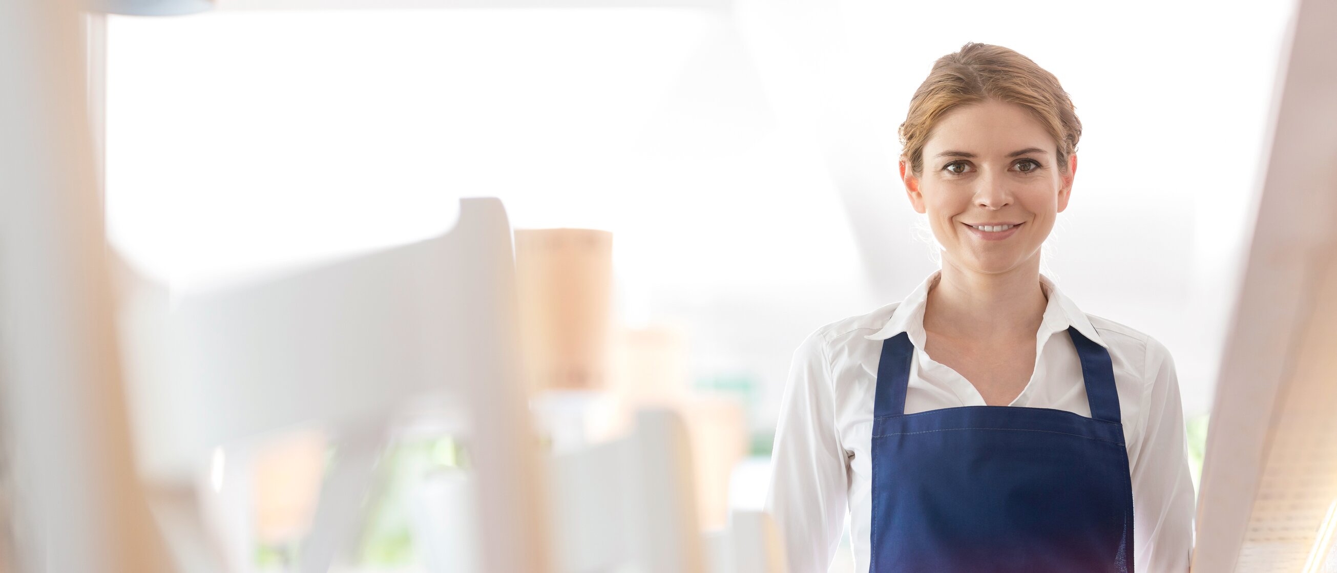 Junge Servicemitarbeiterin im blauen Schürzenoutfit nimmt Bestellungen in einem modernen, hellen Restaurant auf.