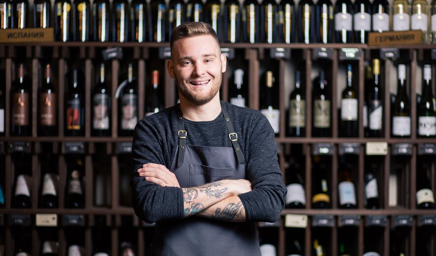 Lächelnder Sommelier mit verschränkten Armen vor einem Weinregal in einem Weinladen.