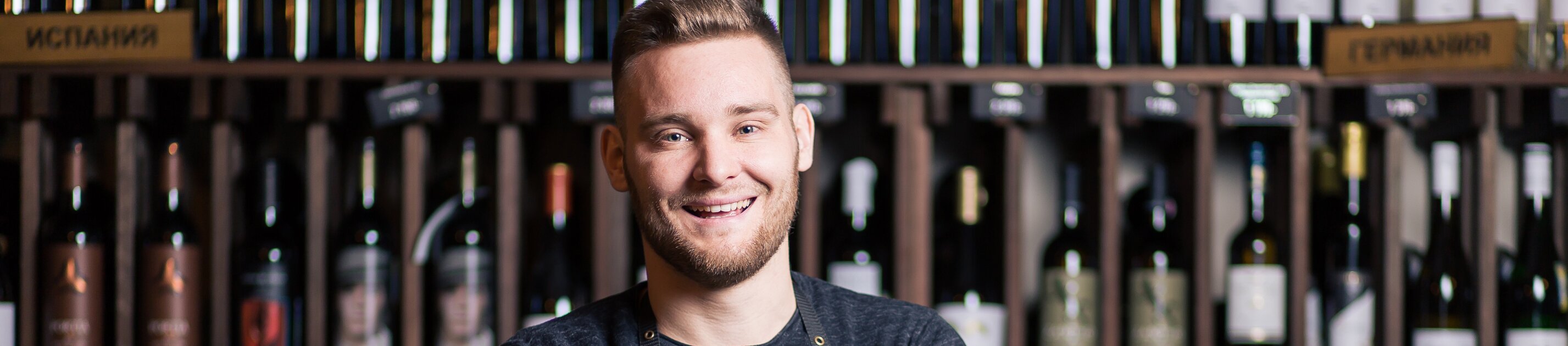 Lächelnder Sommelier mit verschränkten Armen vor einem Weinregal in einem Weinladen.