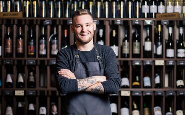 Ein lächelnder Sommelier steht selbstbewusst vor einem Regal voller Weinflaschen und symbolisiert die Professionalität und Leidenschaft, die Teilnehmer in den IHK-Weiterbildungen der Sommellerie erwerben.