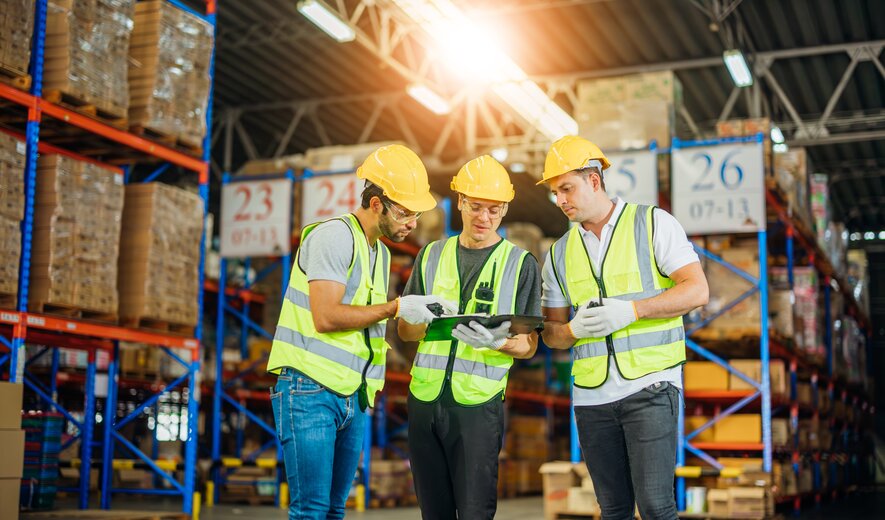 Drei Lagerarbeiter mit Sicherheitswesten und Helmen prüfen gemeinsam ein Tablet in einem großen, gut beleuchteten Warenlager