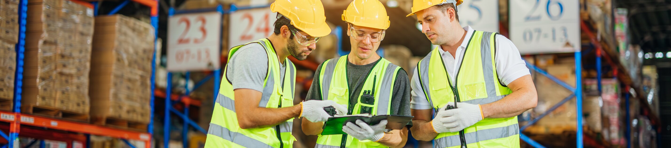 Drei Lagerarbeiter mit Sicherheitswesten und Helmen prüfen gemeinsam ein Tablet in einem großen, gut beleuchteten Warenlager