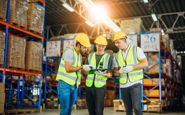 Drei Fachkräfte in Sicherheitskleidung besprechen Warenprozesse in einem Logistiklager – praxisorientierte Weiterbildung zu Import, Export und Zollabwicklung.