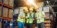 Drei Lagerarbeiter mit Sicherheitswesten und Helmen prüfen gemeinsam ein Tablet in einem großen, gut beleuchteten Warenlager