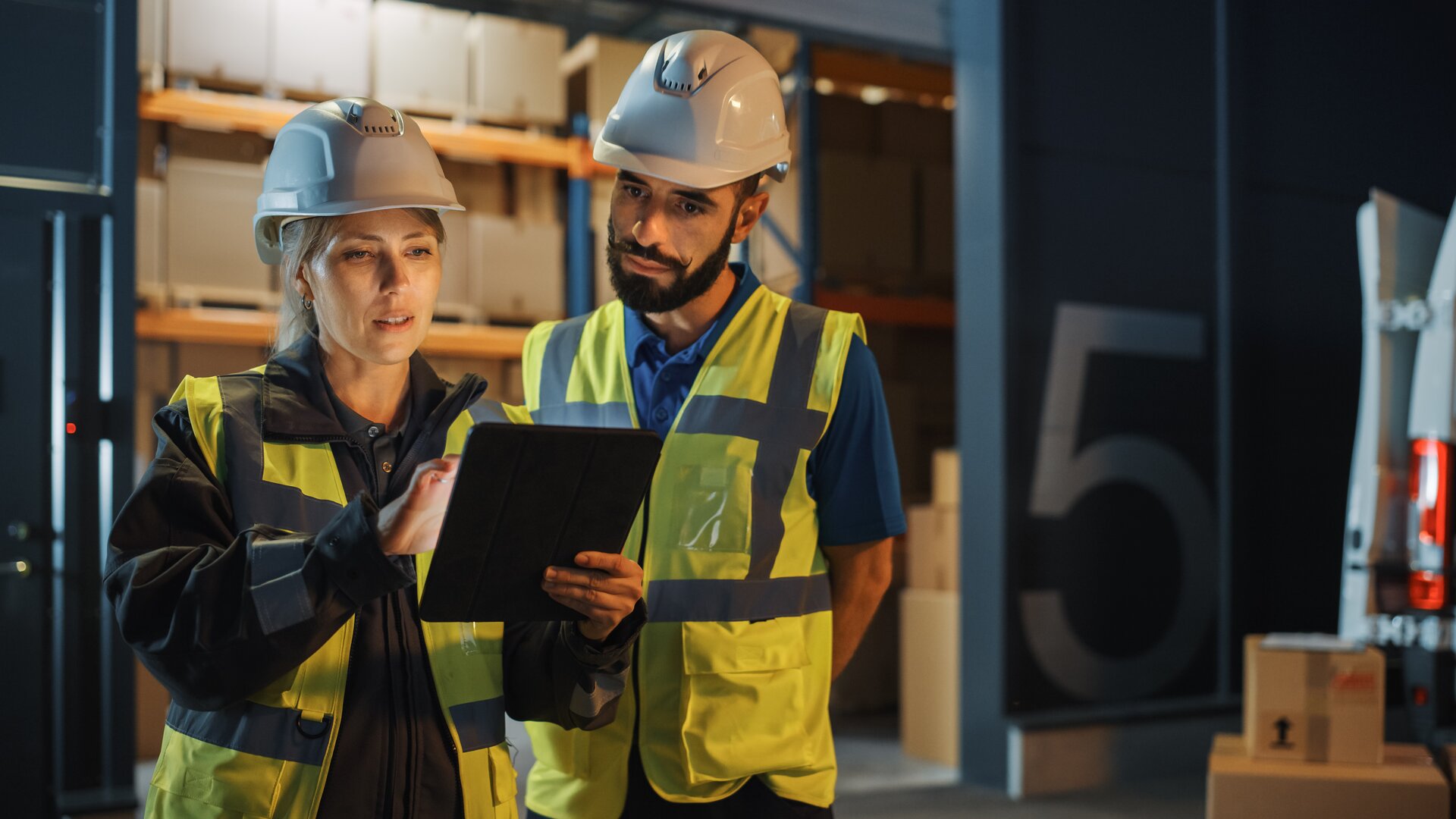 Zwei Lagerarbeiter in Schutzwesten und Helmen überprüfen Daten auf einem Tablet in einer Logistikhalle