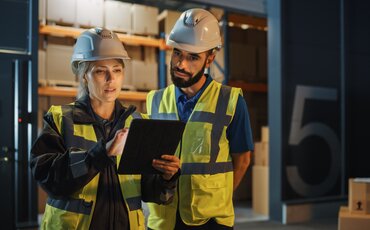 Zwei Mitarbeitende mit Schutzhelmen und Sicherheitswesten prüfen Exportdokumente in einer Lagerhalle – Weiterbildung zur Exportkontrolle und rechtssicheren Außenwirtschaft.