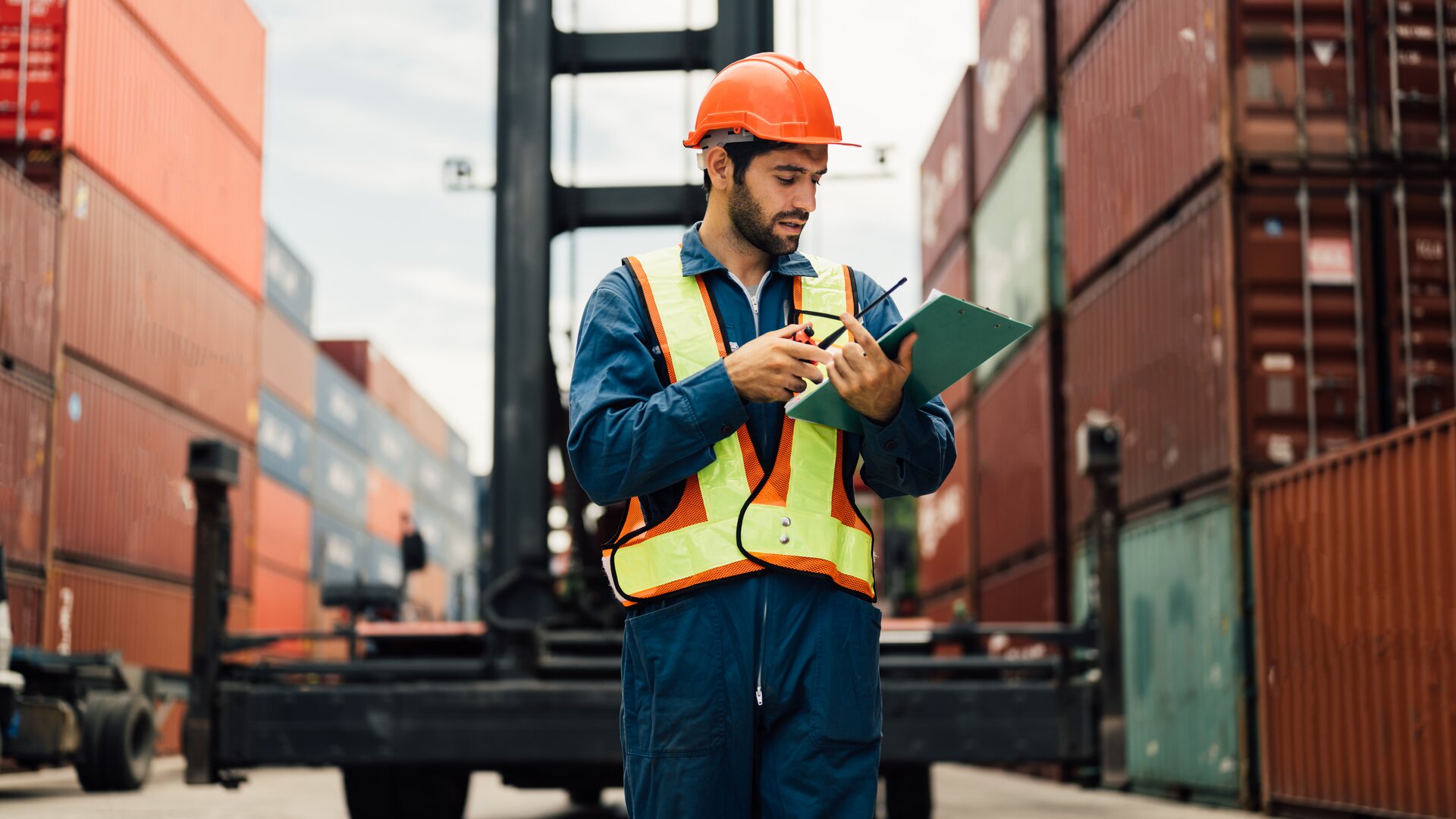 Logistikmitarbeiter mit Schutzhelm und Warnweste prüft Containerdokumente im Hafen zwischen gestapelten Frachtcontainern