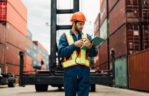 Mitarbeitender in Sicherheitsweste und Helm prüft Dokumente zwischen Containern im Hafen – Weiterbildung zur IHK-Fachkraft Zoll.