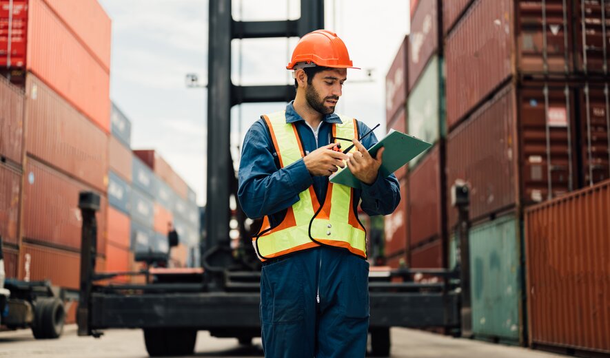 Logistikmitarbeiter mit Schutzhelm und Warnweste prüft Containerdokumente im Hafen zwischen gestapelten Frachtcontainern
