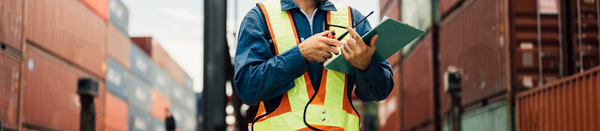 Logistikmitarbeiter mit Schutzhelm und Warnweste prüft Containerdokumente im Hafen zwischen gestapelten Frachtcontainern