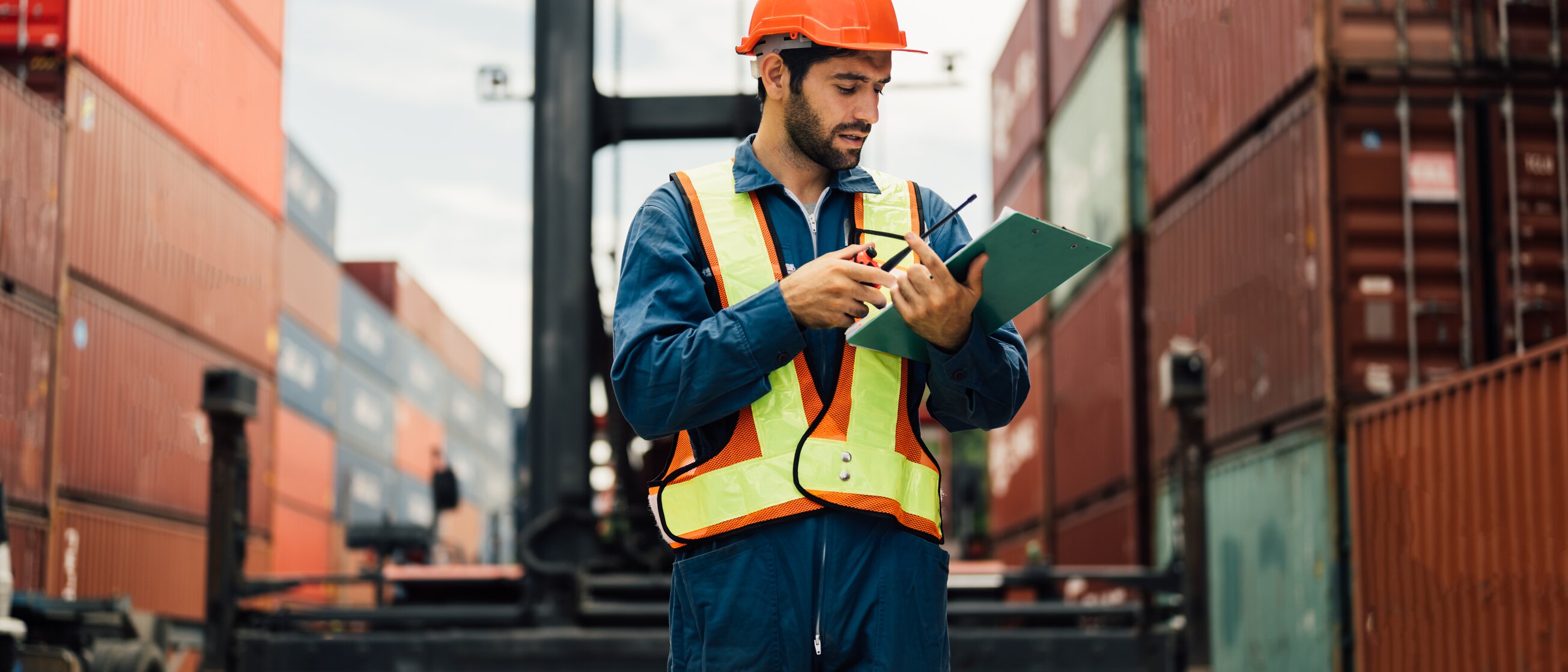 Mitarbeitender in Schutzweste und Helm überprüft Frachtpapiere zwischen Containern – Weiterbildung zur Exportkontrolle und sicheren Zollabwicklung.