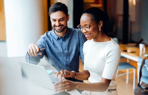 Ein Mann und eine Frau sitzen nebeneinander am Schreibtisch und schauen lächelnd in den Laptop