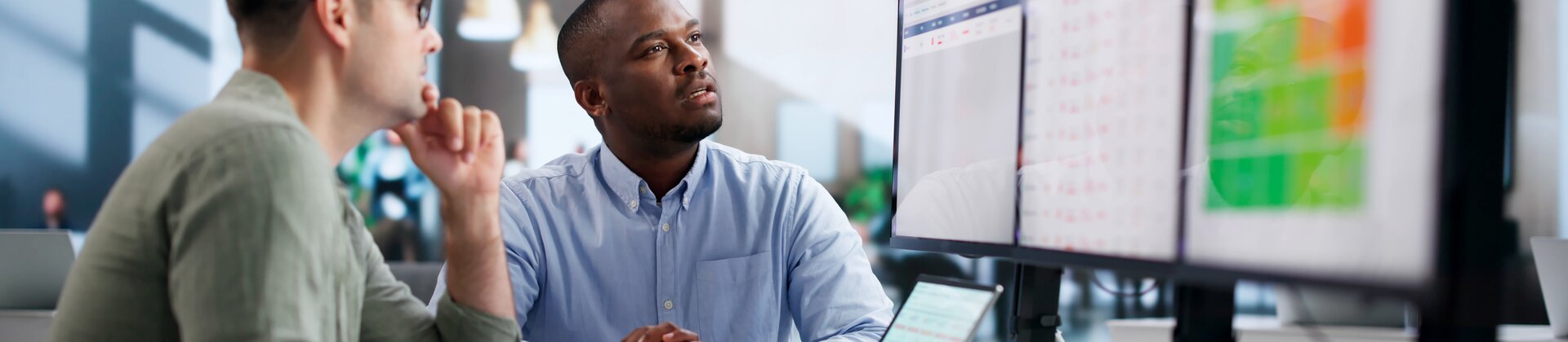 Zwei Mitarbeiter analysieren Geschäftsdaten an mehreren Bildschirmen im Büro.