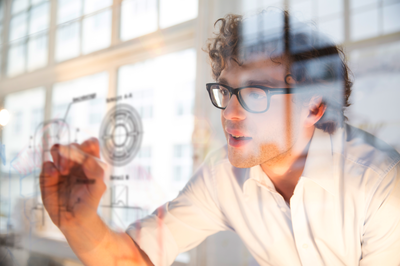 Mann schreibt Formeln auf transparente Glaswand – Symbol für Fachwissen, analytisches Denken und den Bachelor-ähnlichen Abschluss des Industriemeisters Elektrotechnik.