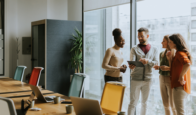 Vier Personen stehen im modernen Büro und besprechen sich mit einem Tablet – Symbol für Coaching, Kommunikation und Teamarbeit in der Ausbildung.