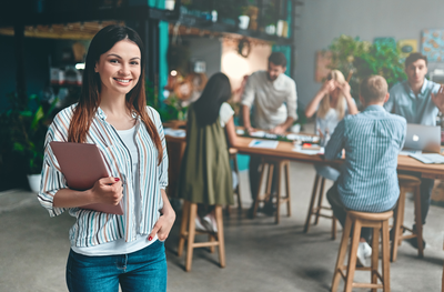 Junge Frau mit Tablet steht in modernem Büro und lächelt – Symbolbild für aktuelle Informationen und Social-Media-Angebote der IHK Akademie Ingolstadt.