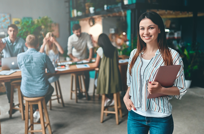 Junge Frau mit Tablet steht lächelnd in modernem Büro – Symbolbild für die Weiterbildung Projektmanager/-in IHK an der IHK Akademie Weilheim