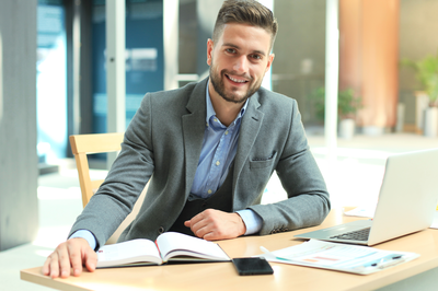 Junger Mann im Anzug sitzt lächelnd am Schreibtisch mit Laptop und Unterlagen – Symbolbild für die Weiterbildung Geprüfte/-r Personalfachkaufmann/-frau