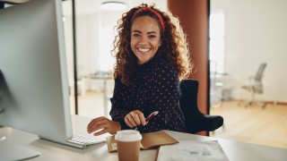Smiling young businesswoman working at her computer and going over paperwork while sitting at a desk in an office