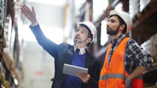 Businessman with touchpad pointing at upper shelf with goods while talking to worker