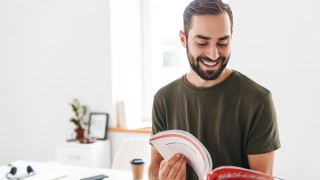 Image of caucasian happy man wearing casual t-shirt reading brochure and smiling while working in bright office