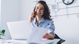 Laughing young brown haired female in glasses in blue shirt multitasking speaking on smartphone and working with notepad in office