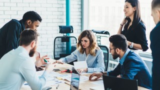 High angle of multiracial business partners in elegant clothes listening to coworker proposition while examining reports and using digital devices at desk in light contemporary conference room