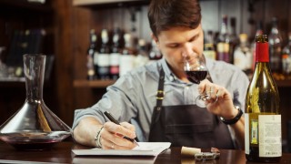 Male sommelier tasting red wine and making notes at bar counter. Bottle of wine nearby. Professional expert appreciates quality of alcoholic beverage, degustation process