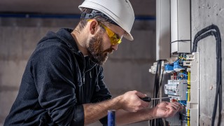 A male electrician works in a switchboard with an electrical connecting cable, connects the equipment with tools.