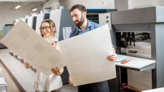 Young woman designer and print operator working with paper print at the print manufacturing with offset machine on the background